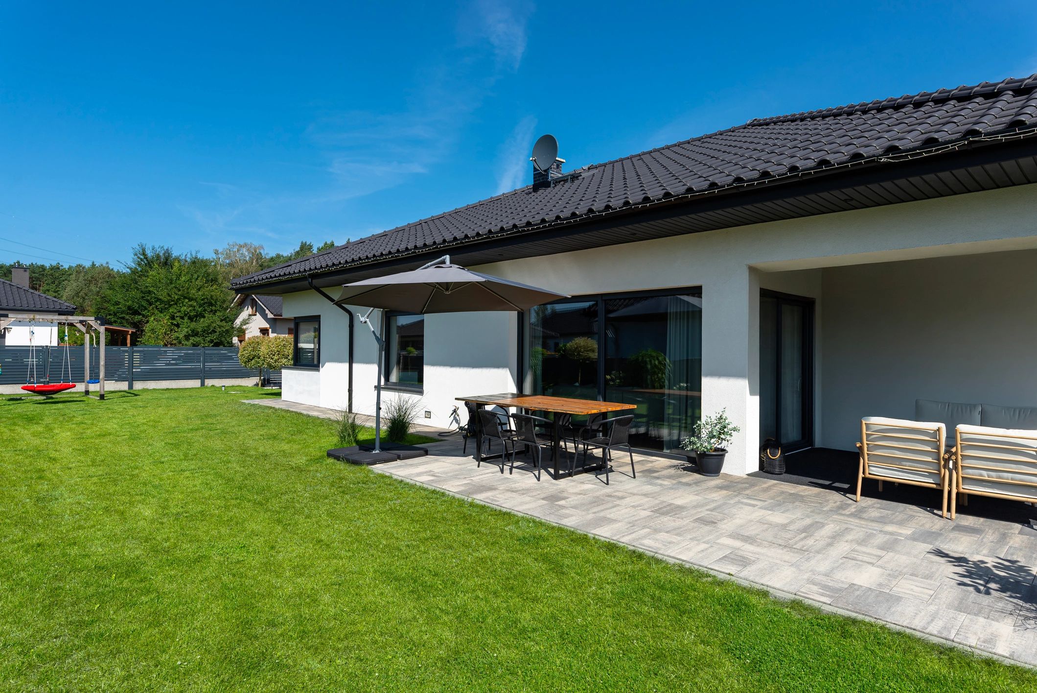A beautiful garden with green grass, a patio table and umbrella visible, a house in beautiful summer weather, a very wide shot.